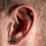 Close-up van een oor met meerdere piercings in de oorlel en het kraakbeen, waaronder een groene steen