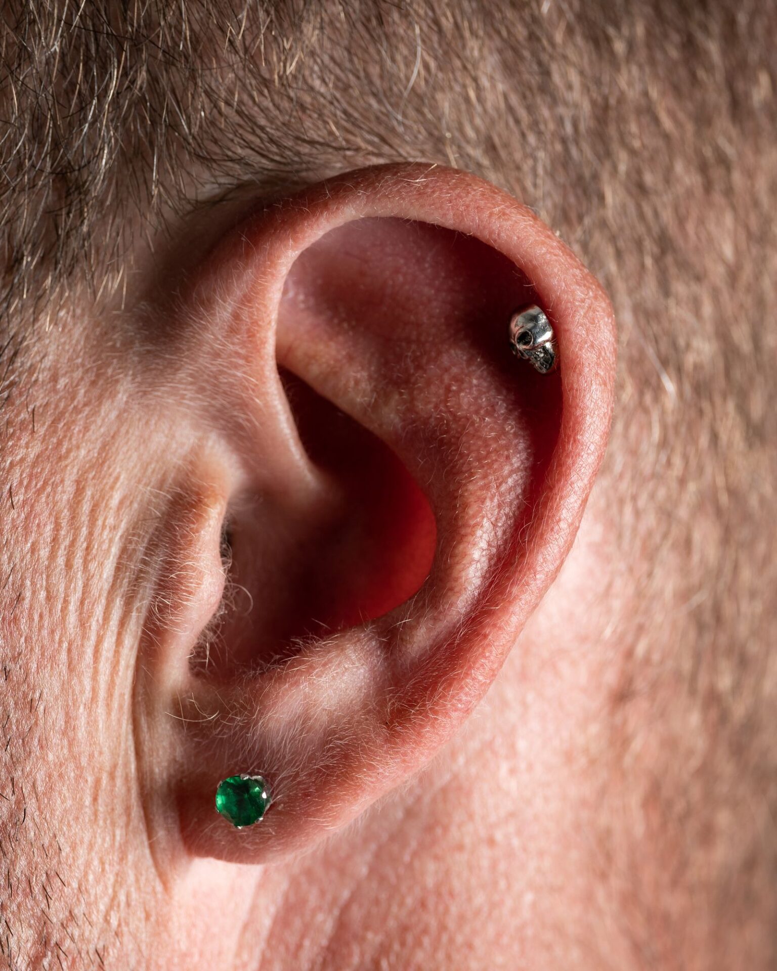 Close-up van een oor met meerdere piercings in de oorlel en het kraakbeen, waaronder een groene steen